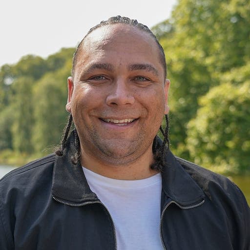 Alex Holmes OBE, Trustee of the Queen Elizabeth Trust, smiling at the camera in an outdoor portrait in a leafy parkland setting.