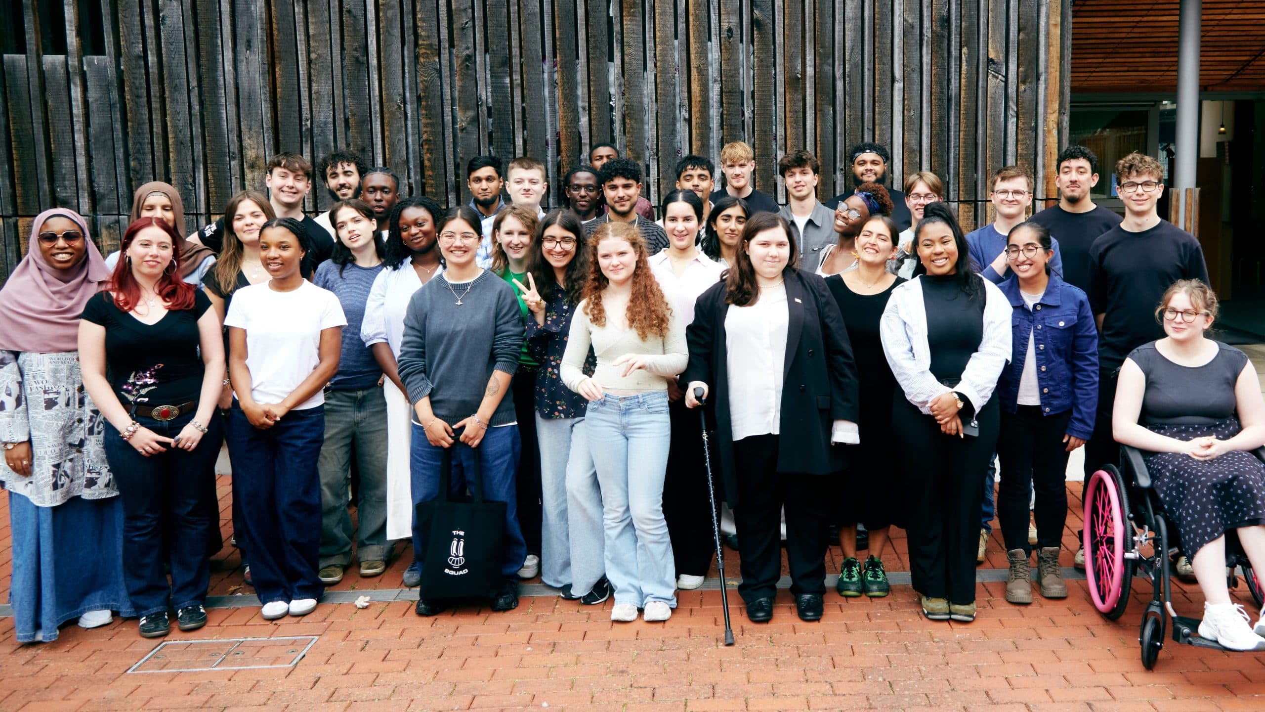 Around 40 young people from My Life My Say gathered for a group portrait outside a timber-clad building, standing on a brick courtyard.