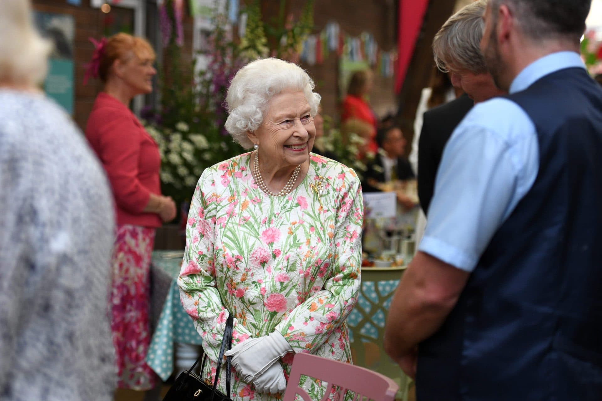 Queen Elizabeth smiles while greeting guests at an outdoor event, wearing a pink and green floral dress and pearls.