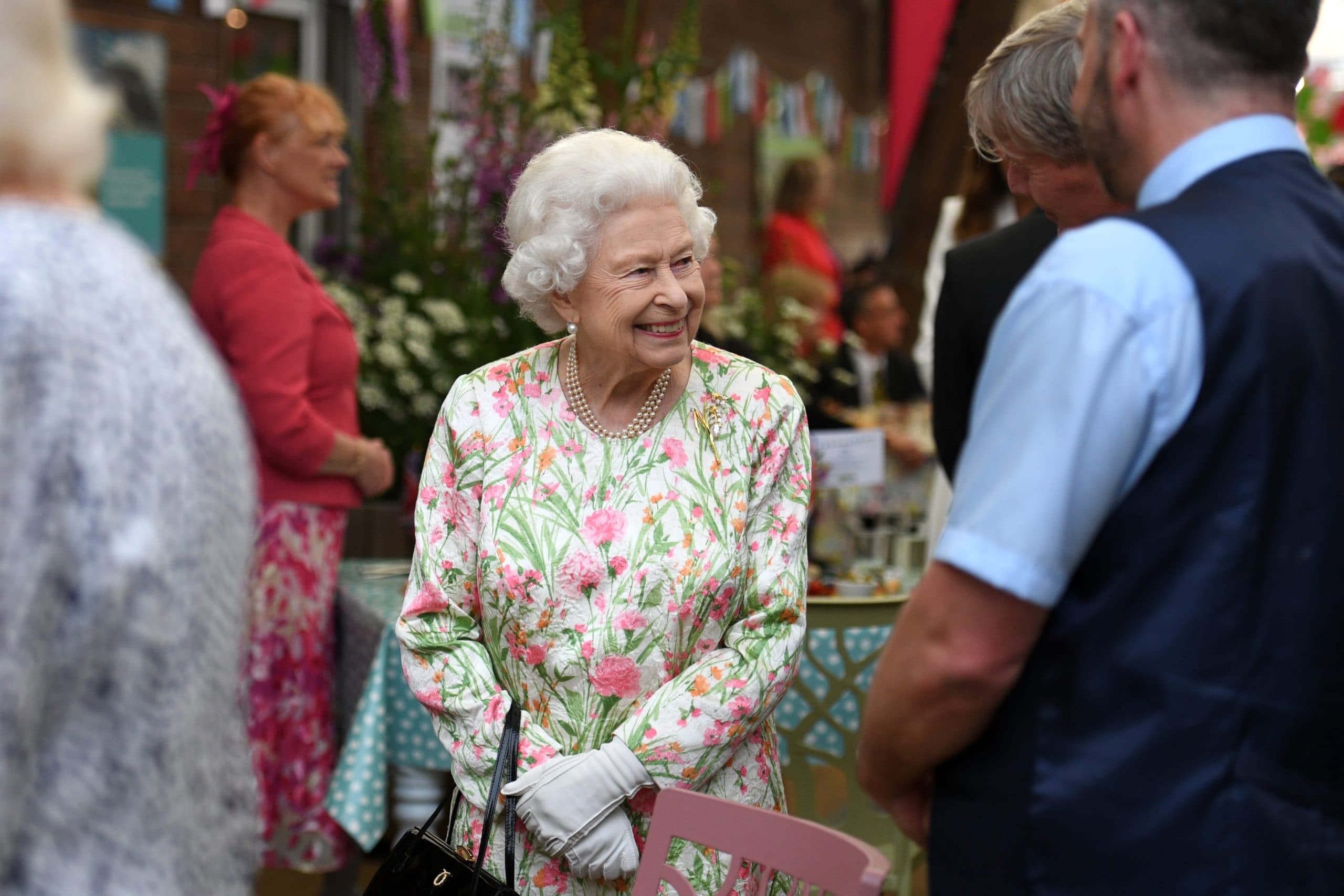 Queen Elizabeth smiles while greeting guests at an outdoor event, wearing a pink and green floral dress and pearls.