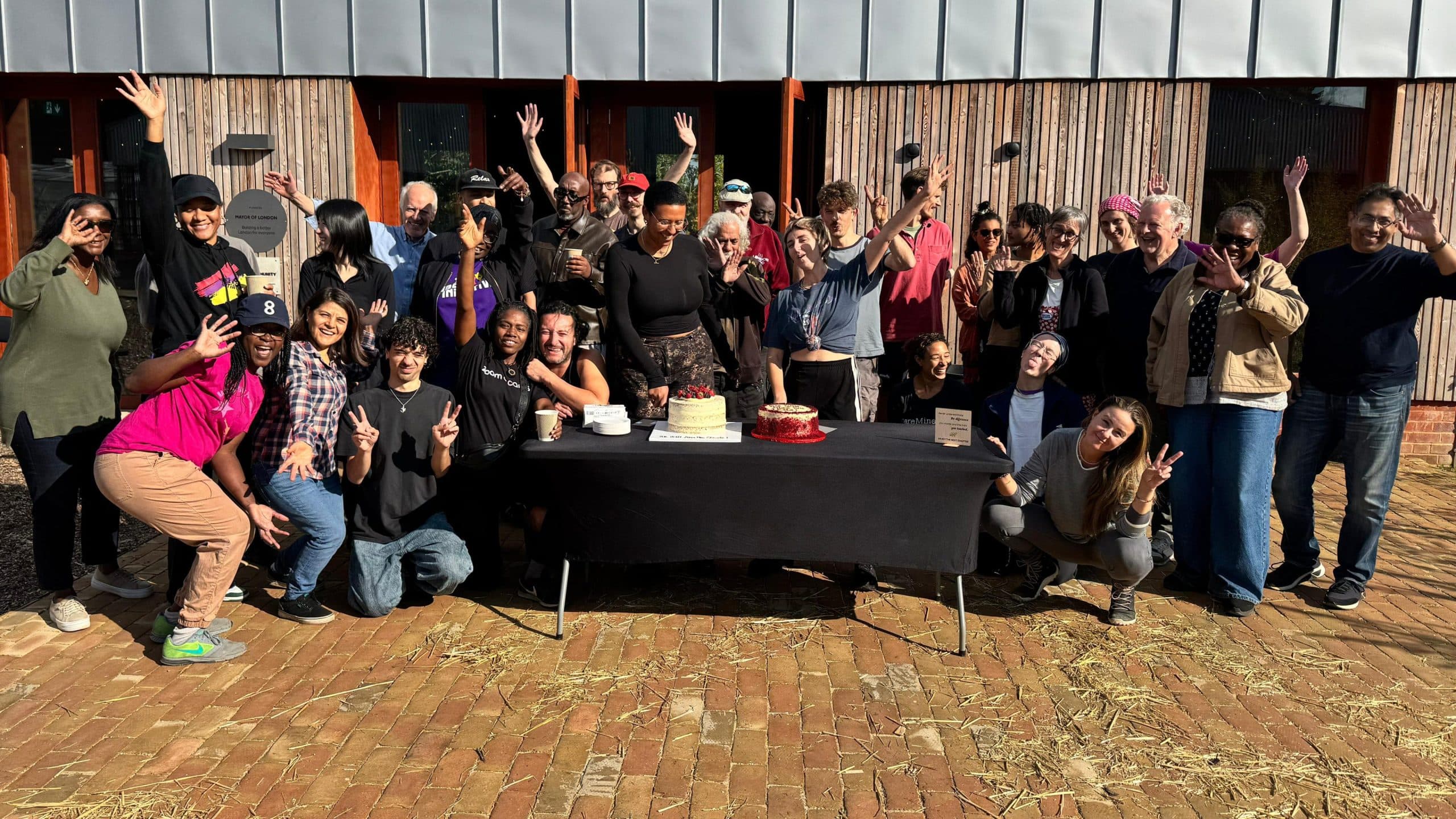 Members of The Ubele Initiative gathered outside a timber-clad community building on a sunny day, waving and posing around a table set with two celebration cakes.