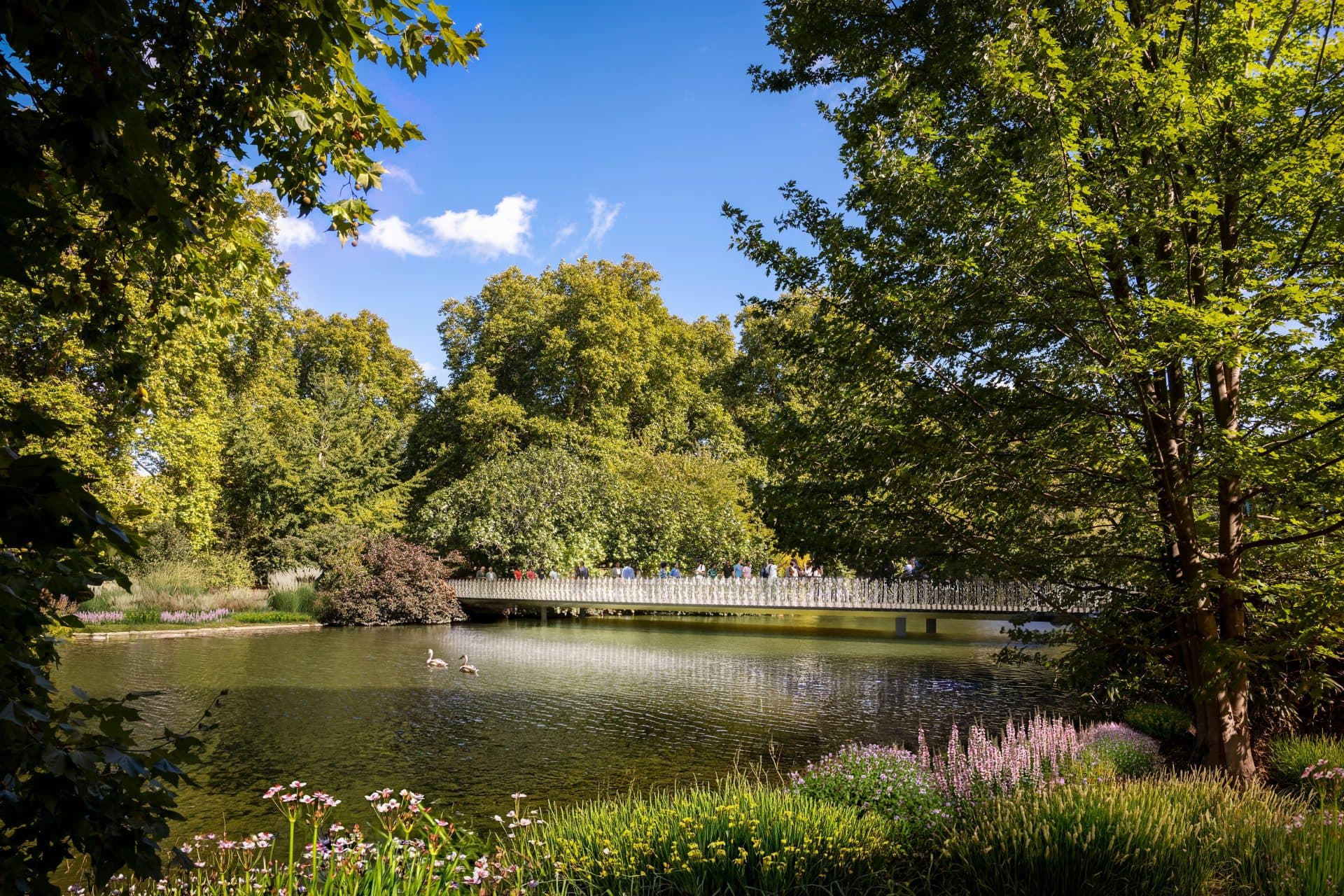 National Memorial Foster and Partners. A sunlit pedestrian bridge with a translucent balustrade spans a tree-lined lake in St James's Park, with swans on the water and visitors crossing.